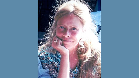 A middle aged white woman with blond hair wearing a multicolored top smiles a closed mouth smile at the camera with her head propped on her hand