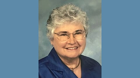 Older white woman with short silver and white hair wearing a blue top, cross necklace and wire glasses smiles warmly