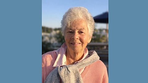 Older white woman with short white hair wears a pink sweater with a grey sweatshirt tied around her shoulders