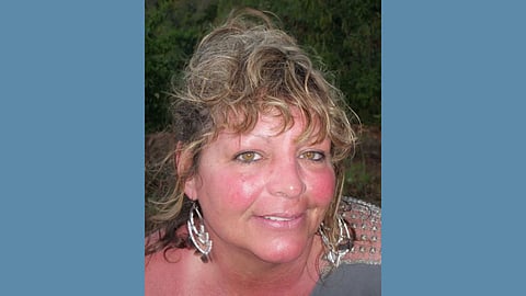 A middle aged white woman with wispy dark blonde hair wearing long dangling earrings and smiling at the camera