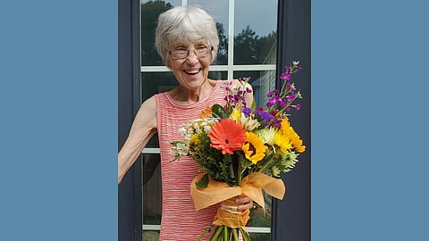 Elderly white woman with short white hair is wearing a orange and white striped tank top and holding a bouquet of flowers