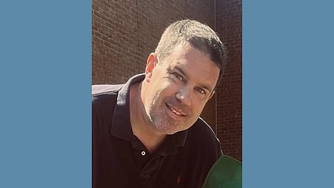 Middle aged white man in a black polo with close cropped brown hair and stubble smiles at the camera