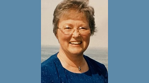 An older white woman with short grey hair wears glasses and a navy dress and jacket set and smiles at the camera