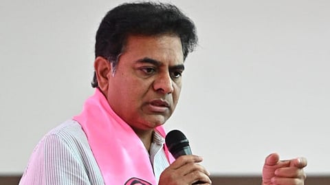 A medium close-up photograph shows K. T. Rama Rao, an Indian politician, wearing a white striped shirt and a pink BRS (Bharat Rashtra Samithi) party scarf with images of K. Chandrashekar Rao (KCR) and an outline of India on it. He is speaking passionately into a black microphone held in his right hand, gesturing with his left hand towards an unseen audience. He is indoors, likely at a party meeting or rally, with a plain white wall visible behind him. His expression is serious and engaged.