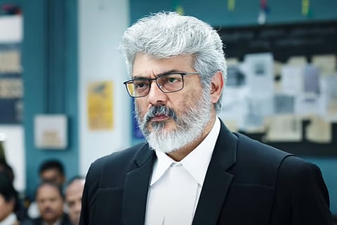A trailer screengrab of Ajith Kumar in black lawyer's robe, glasses, and a beard. Other people can be seen behind him. The walls are white and blue with a large notice board.