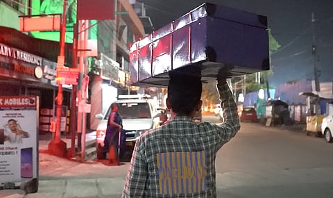 Goura Naik, wearing a plaid shirt and carrying a luggage case on his head, walks towards the Ernakulam Railway Station as the camera captures him from behind.