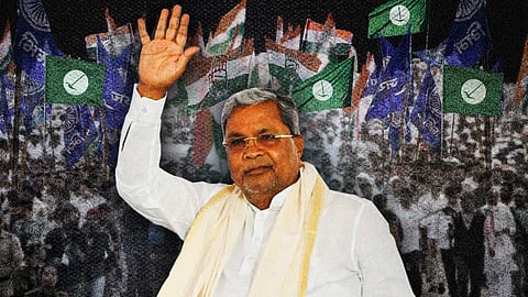 Karnataka Chief Minister and Congress leader Siddaramaiah, dressed in a white traditional kurta and shawl, waves to a large crowd. The background shows numerous flags from political parties, including Congress flags, against a densely packed crowd, creating the atmosphere of a political rally or public event.