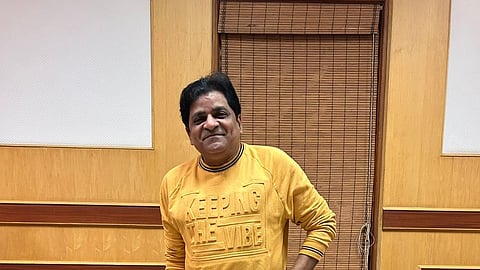 Telugu actor Ali in a yellow sweatshirt which says 'Keeping the Vibe' stands in a room with brown leather couches and wood-paneled walls. He is wearing light jeans and gray sneakers, smiling at the camera.