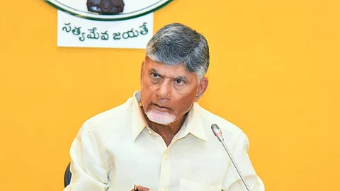 Andhra Pradesh Chief Minister Nara Chandrababu Naidu in a light yellow shirt sits at a desk, speaking into a microphone. He holds a pen and has documents in front of him. Behind him is the Andhra Pradesh government's emblem on a yellow wall.