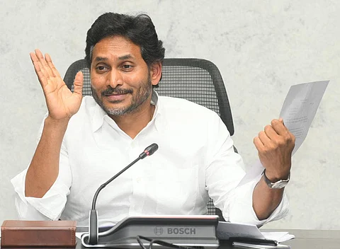 YSRCP chief and former Andhra Pradesh Chief Minister YS Jagan Mohan Reddy in a white shirt sits at a desk, holding a piece of paper in his right hand and gesturing with his left. A microphone is in front of him, and a Bosch branded panel is seen on the table.