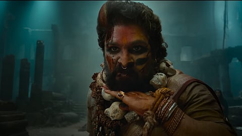 Allu Arjun in Pushpa: The Rule wearing a maroon saree and a garland of skulls, with his face covered in vermilion. He is running the back of his palm along his chin, with a dilapidated building in the background.