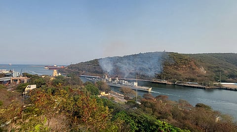 A scenic view of a coastal area featuring a harbor with docked ships and cranes. Smoke rises from an industrial area surrounded by lush green hills. The calm sea stretches into the horizon under a clear sky.