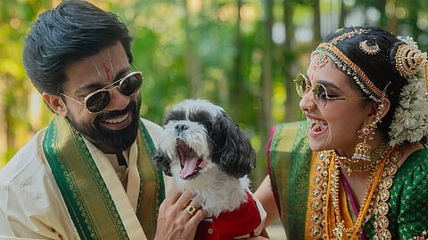 Keerthy Suresh and Antony Thattil in wedding attire laugh while holding a small black and white dog wearing a red outfit. They are outdoors with greenery in the background.