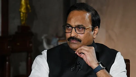 TV9 Managing Editor Rajinikanth with glasses and a mustache sits thoughtfully in a chair with lion-shaped armrests. He is wearing a black vest over a white shirt and holding papers. The background is dimly lit with a statue visible on a table.