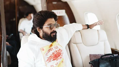 Allu Arjun in a white hoodie sitting inside an aircraft. His left arm is on the empty seat next to him, and there's a phone on the table in front of him. He is wearing sunglasses.