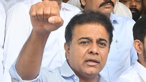 BRS leader KTR in a blue striped shirt raising his fist. There are a group of people behind him standing against a wood-panelled wall.