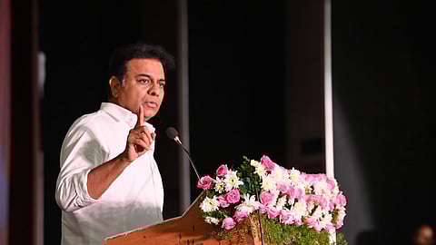 BRS leader KTR in a white shirt speaks at a podium adorned with pink and white flowers. He gestures with one hand while addressing an audience.