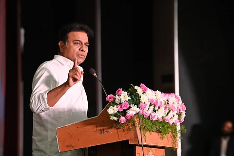BRS leader KTR in a white shirt speaks at a podium adorned with pink and white flowers. He gestures with one hand while addressing an audience.
