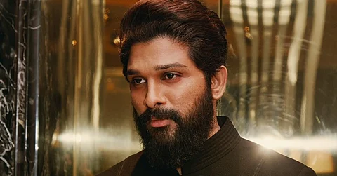Allu Arjun in a black suit staring intensely to his left, standing against a shiny backdrop