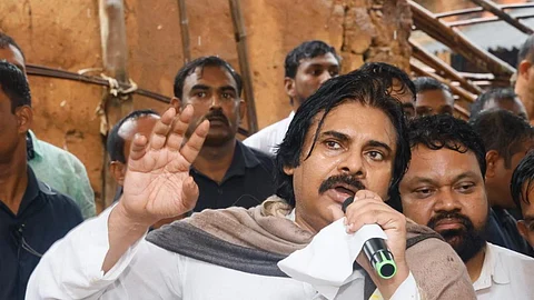 Pawan Kalyan speaking into a mic during a visit to Bagujolu village. He is wearing a white kurta and a brown shawl, with his right hand raised and holding a mic with his left. There's a mud wall in the background and a few villages and security staff around him.