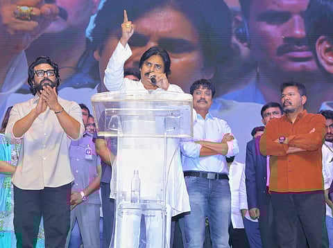 Actors Ram Charan, Pawan Kalyan, SJ Suryah and director Shankar on a stage. Pawan Kaylan is speaking into a mic asertively while raising his right hand with the index finger pointing up, Ram Charan is on his right clapping, SJ Suryah and Shankar are on his right with folded arms.