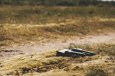 A black revolver, symbolising Maoist surrender, rests on the grass in an outdoor setting, with a blurred background of vegetation and a dirt path.