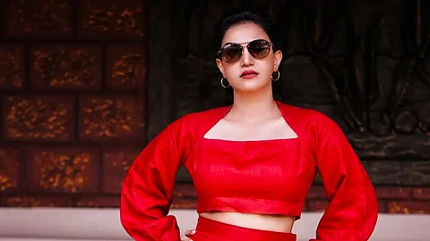 Honey Rose in a red outfit, gold hoop earrings, and sunglasses, posing confidently with hands on hips.