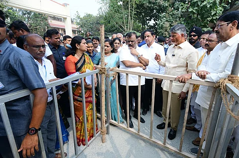 Andhra Pradesh Chief Minister Chandrababu Naidu in off-white shirt and trousers standing next to a barricade. With his left hand on the barricade, he is addressing officials and Ministers. He is surrounded by many men and women.