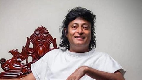 Portrait of Boby Chemmanur seated on an ornate wooden chair, wearing a white outfit and smiling.