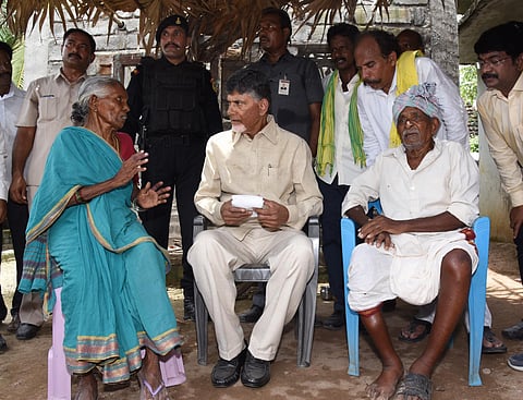 Andhra Pradesh Chief Minister Chandrababu Naidu is seated in a chair. An old woman is seated on a chair to his right, and an old man on his left. The woman is wearing a blue saree and talking to Naidu. They are surrounded by security personnel and others, and are sitting under a thatched roof.