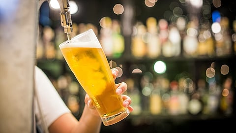 A glass of beer with Heineken imprinted on it is being filled from a tap by a person whose arm is visible. They are wearing a white t shirt. There are shiny alcohol bottles blurred in the background.