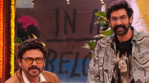 Actor Venkatesh is sitting on a couch while his nephew Rana Daggubati is leaning on the couch standing next to him. They are on a studio set. Venkatesh is wearing a white shirt, white pancha and brown jacket. Rana is wearing a black t shirt with Bob Marley's image and and a black and white overshirt, and a pancha. They are both wearing sunglasses and smiling at the camera.