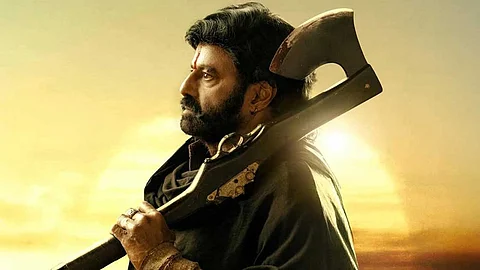 Actor Balakrishna with a beard and intense expression holds a large axe over his shoulder, set against the backdrop of a glowing sunset. He is dressed in dark clothing, with the sunlight emphasizing his profile.