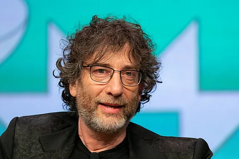 Author Neil Gaiman wearing a beard, glasses and black clothes, seated in front of greenish-blue and white background.