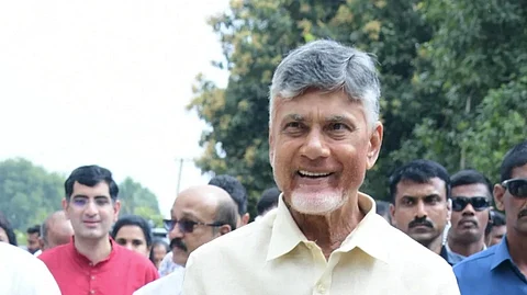 Andhra Pradesh Chief Minister Chandrababu Naidu dressed in a cream-colored shirt and traditional white attire is smiling while walking outdoors. He is surrounded by a group of people, some in casual traditional attire and others in formal security uniforms. Trees and greenery are visible in the background, suggesting a public or cultural event.