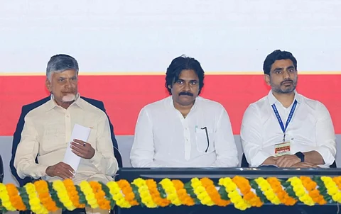 The image shows three individuals seated in a row at an event - Andhra Pradesh Chief Minister Chandrababu Naidu, Deputy CM Pawan Kalyan, and Minister Nara Lokesh. They are dressed in traditional or formal attire, with a floral garland arrangement in the foreground. The backdrop includes a plain, colored panel.