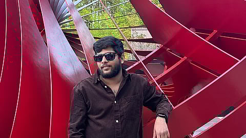 Ravi Teja wearing a dark brown button-up shirt and white trousers is casually leaning against a red structure with curved and angular shapes. He has sunglasses on and a relaxed expression, standing on a paved surface with lush green trees visible in the background.
