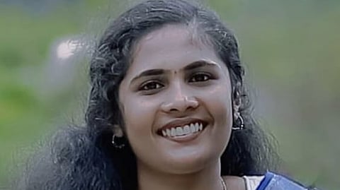 Sruthi, wearing a white and blue saree, standing outdoors with greenery in the background