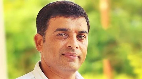 The image shows Telugu producer Dil Raju with a warm and calm demeanor. He has short black hair with a slight salt-and-pepper tone, neatly combed, and is wearing a white shirt. His expression is gentle, with a faint smile. The background is softly blurred with greenery, giving the image a bright and natural feel. The lighting is soft and evenly distributed, highlighting his face without any harsh shadows.