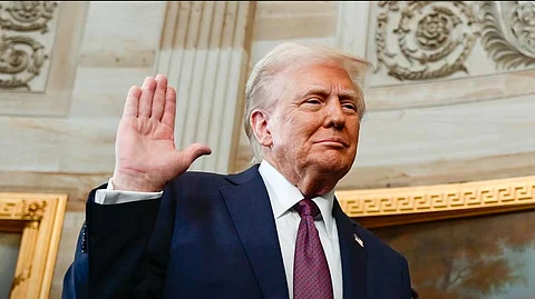 Donald D Trump with his right hand raise, facing outwards, while taking oath as US President. He is wearing a dark suit, white shirt and red tie. Paintings with gilt frame and ornate architecture can be seen behind him.
