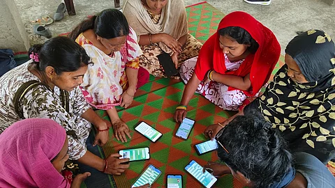 Eight women sitting on the floor in a circle with a mobile phone each in front on them on a mat, participating in a digital literacy training programme.