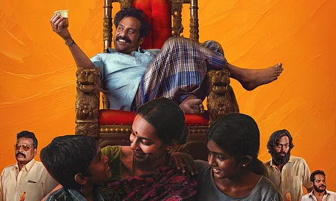 Actor Guru Somasundaram sits on a high-backed wooden chair with red upholstery. He is wearing a light blue shirt and lungi, holding a glass of liqour while dangling his leg over the side. Actors Sanchana and two child actors are in the foreground. The background is bright orange.