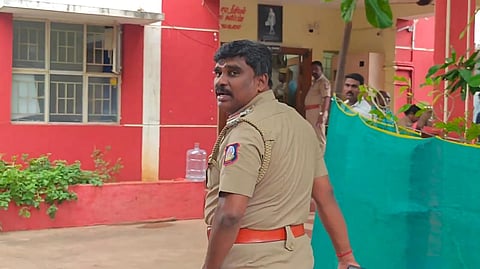 Inspector Gunasekaran is in khaki uniform, turning to look back while walking towards the police station. The station is painted bright red and decorated with potted plants. Several other police personnel can also be seen in the background.