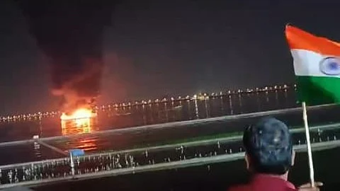 The image shows a night scene where a fire is blazing on a body of of the Hussain Sagar lake. A plume of dark smoke rises from the fire. In the foreground, there are spectators, one of whom is holding an Indian flag.
