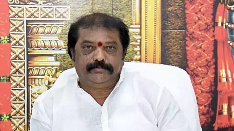 The image shows Gummanur Jayaram seated, wearing a white shirt, with a red mark (tilak) on his forehead. Behind him is a colorful background featuring what appears to be traditional Hindu temple architecture. He is resting his hands on a desk with some papers and a mobile phone placed on it.