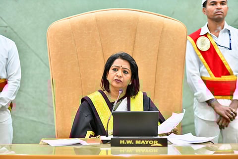 This image features GHMC Mayor Gadwal Vijayalaxmi seated at a desk labeled "H.W. Mayor." She is presiding over a Greater Hyderabad Municipal Corporation council meeting, wearing official attire with a gold and black sash. The background includes two individuals dressed in ceremonial uniforms, adding to the formal setting.