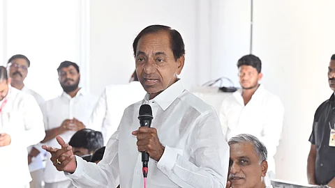 The image shows former Telangana Chief Minister KCR in a white traditional Indian outfit speaking into a microphone while gesturing with his hand. He is surrounded by several people, also dressed in white, some of whom are seated and clapping. The setting appears to be a political gathering indoors, with papers and a table visible in the foreground.