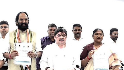 A group of people, including politicians and officials, standing behind a table at a press conference or official event. Some individuals are holding documents with government or official insignia. The central figure, wearing a scarf with political party colors, holds a booklet. The background consists of a plain white wall, and microphones are placed on the table in front of them.