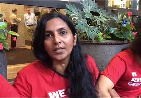 Former Seattle city council member Kshama Sawant, in red a t-shirt, speaking on camera. Plastic potted plants, an shop windows with mannequins can be seen in the background.