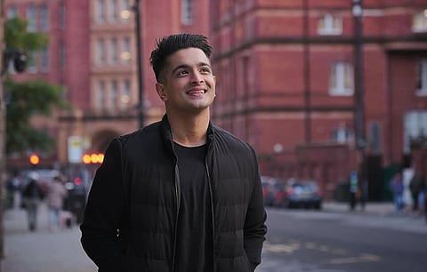 YouTuber Ranveer Allahbadia (alias BeerBiceps) in a black T-shirt and black jacket, smiling in front of a red brick building. The lights and traffic in the background is visible, but blurry.